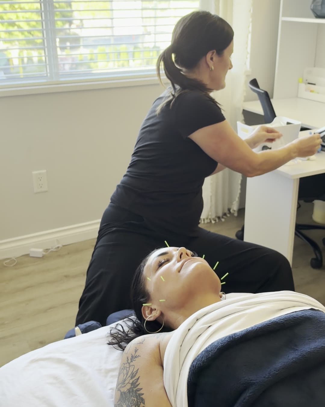 Fine acupuncture needles placed while a client rests face up, with the practitioner at the desk nearby